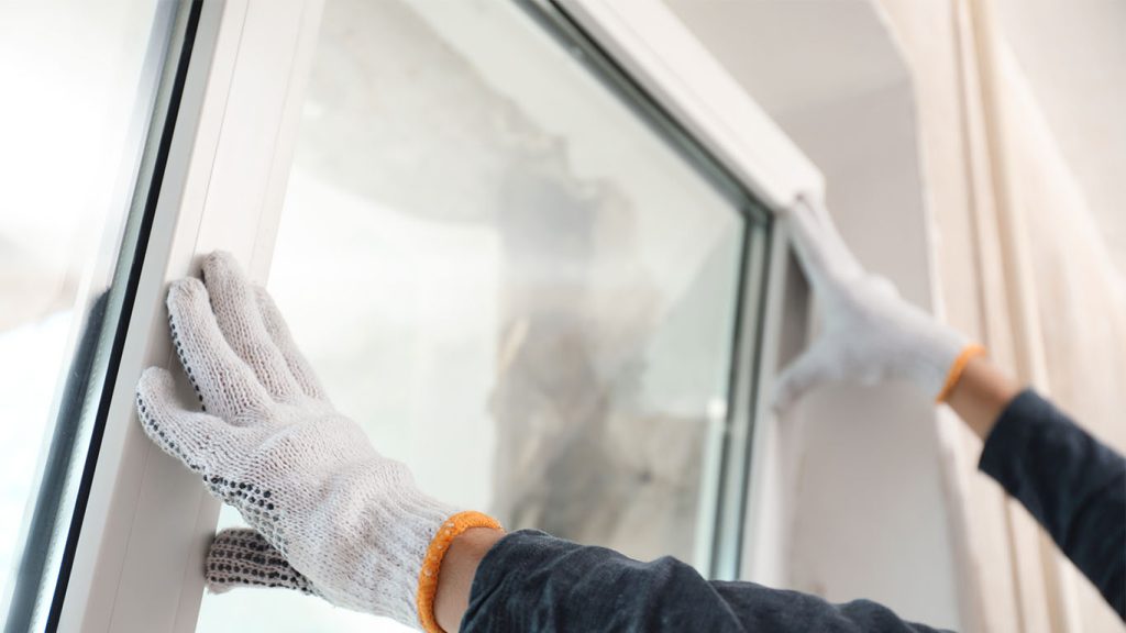 Man in white overalls and cap painting a wall bright orange with a roller, tools on belt, paint bucket nearby, sunlight from window.