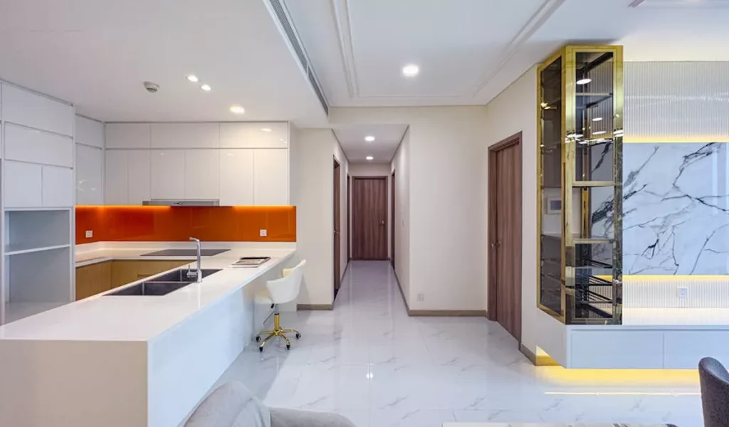 Modern minimalist kitchen with white cabinets, orange backsplash, marble floors, hallway, wooden doors, and a glass display cabinet with gold accents and lighting.